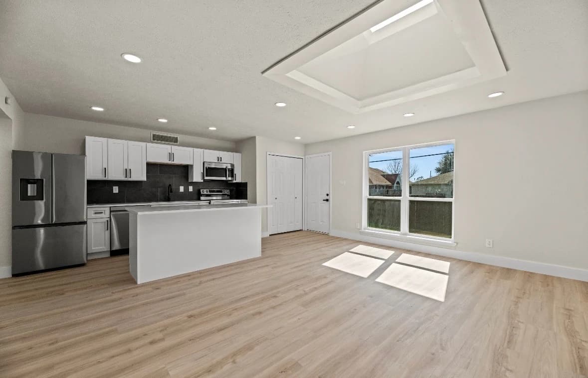 Open living area with wood-look flooring leading to a white kitchen island and stainless refrigerator