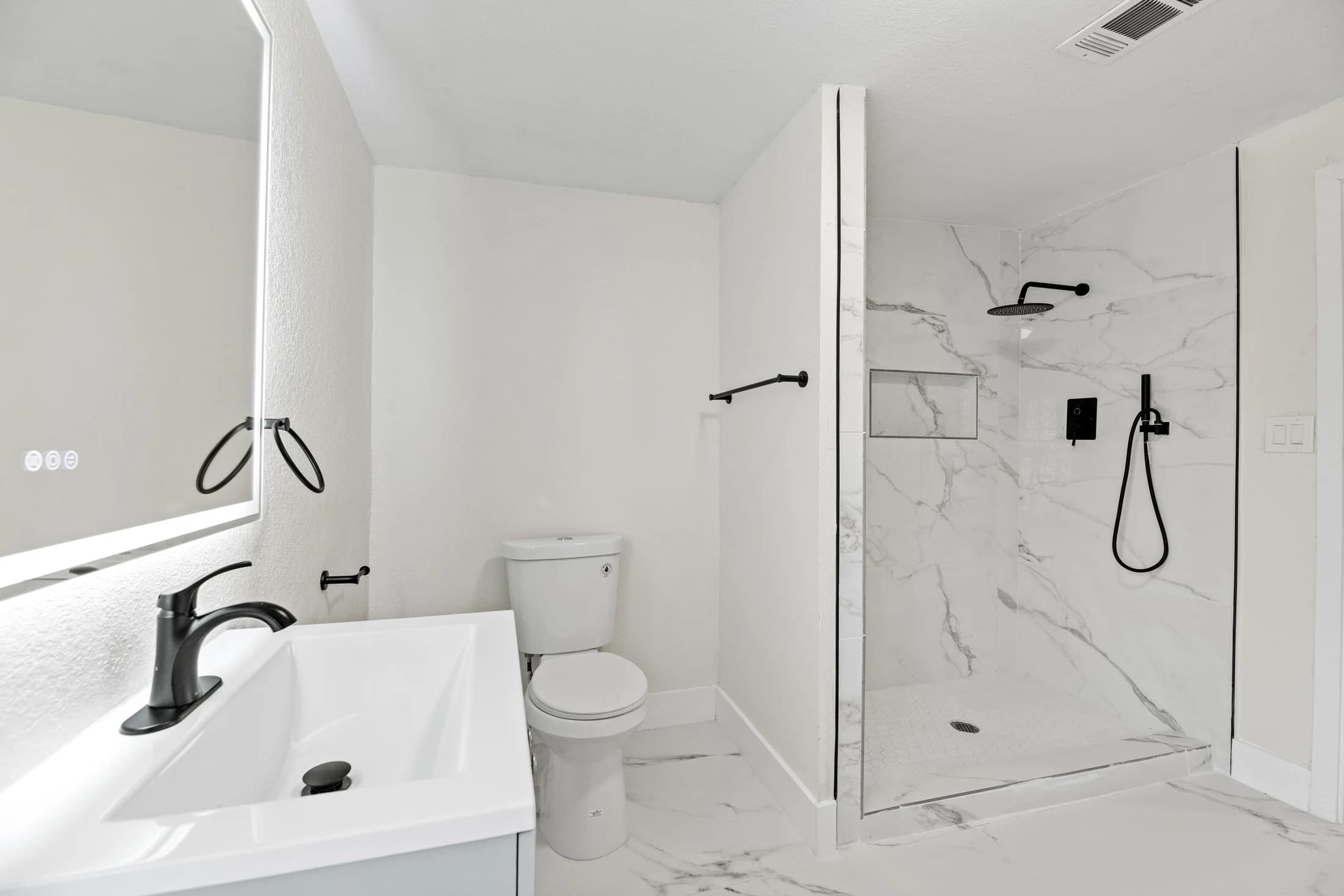 White bathroom vanity, illuminated mirror and glass shower with veined wall tile and black fixtures