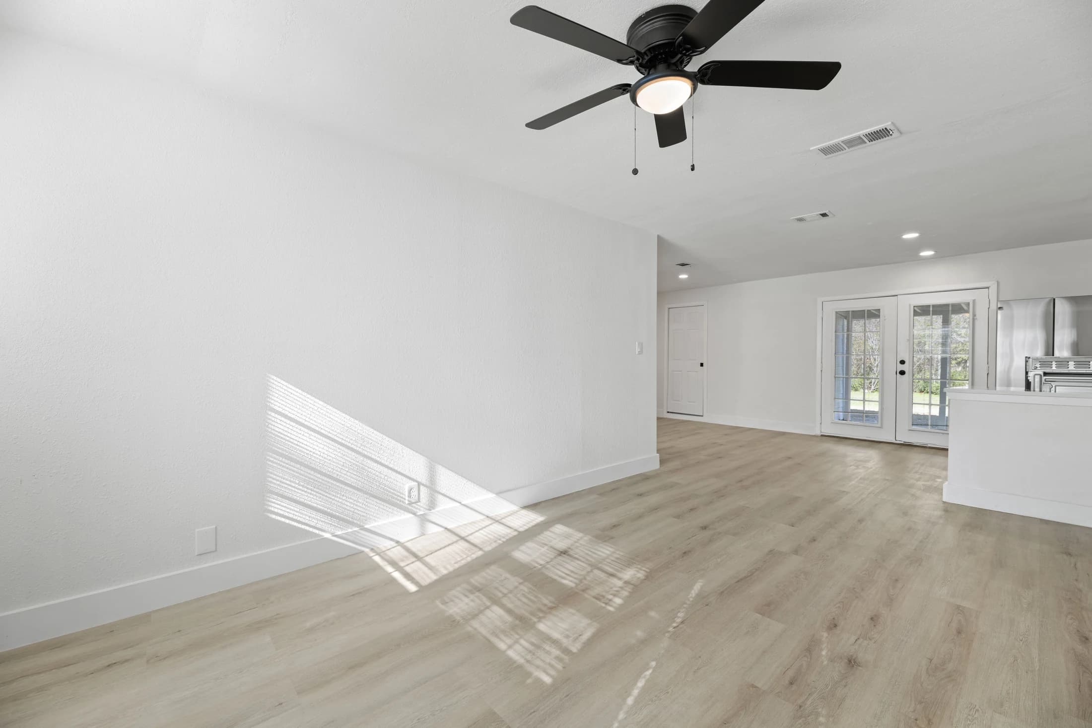 Pale wood-look plank flooring through an empty living room toward glass double doors and the kitchen