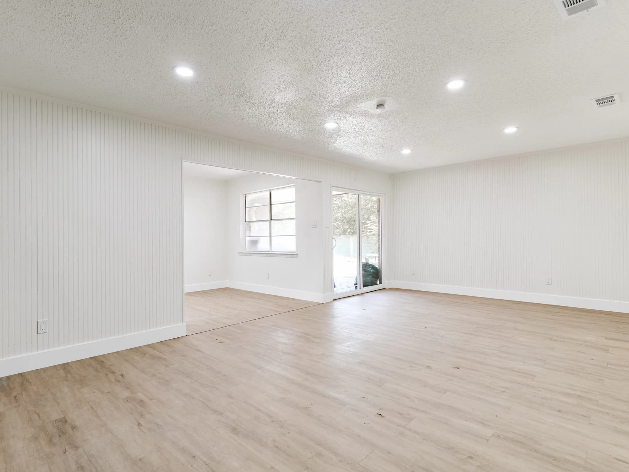 Empty living area with pale plank flooring, white walls, recessed lighting and a view into the next room
