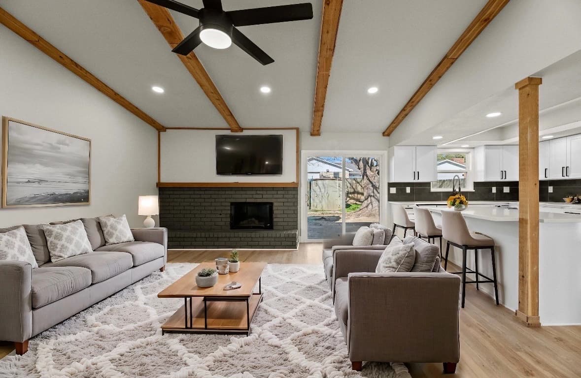 Finished living room with dark brick fireplace, timber beams and an open connection to the white kitchen