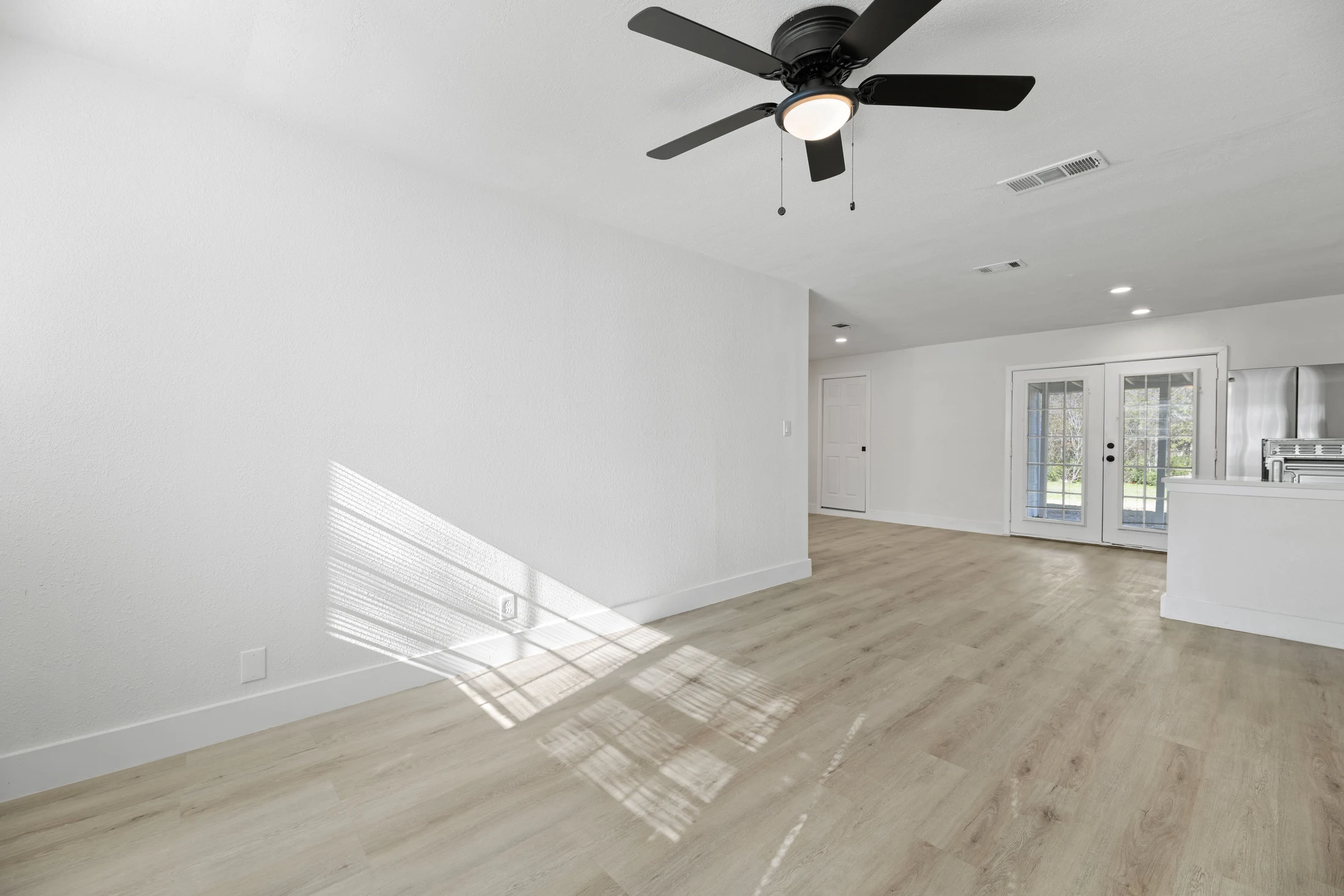 Pale wood-look plank flooring through an empty living room toward glass double doors and the kitchen